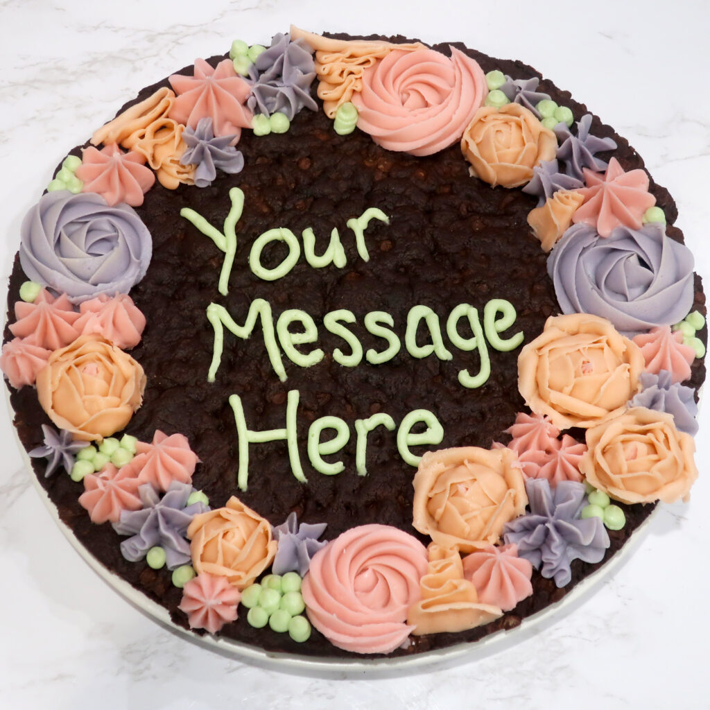 Double chocolate cookie cake with pastel colored buttercream roses and textured piping around the circumference. "Your Message Here" piped in buttercream in the center. These treats are a perfect alternative to cake!