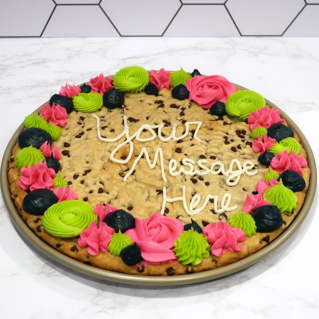 Chocolate Chip cookie cake decorated in pink, green & navy textured buttercream piping around the circumference. the text "Your Message Here" piped in white buttercream in the center. These treats are a perfect alternative to cake!