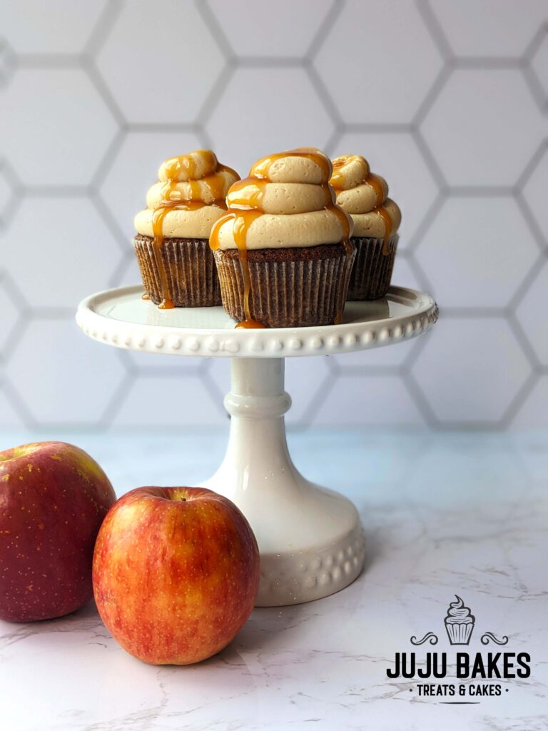 Three apple spice cupcakes with caramel frosting and a caramel drizzle sit atop a white cake stand. Next to the cake stand are two gala apples.