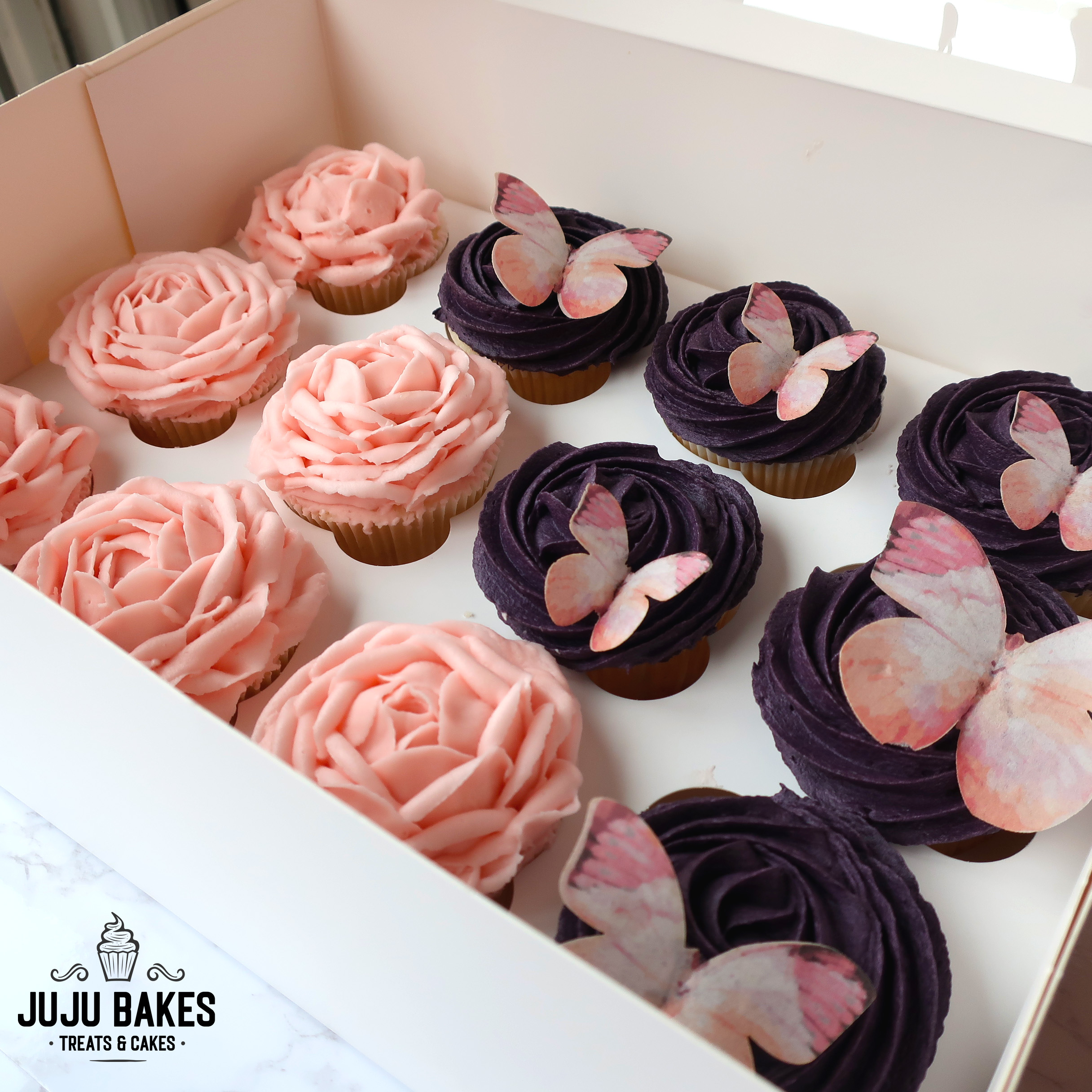 Cupcake box filled with cupcakes decorated in peach-pink and dark purple buttercream flowers and pink butterflies