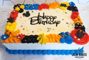 Mickey Mouse themed sheet cake. White cake with piped blue bottom border and blue, orange, red, yellow and black textured piped top border. Black fondant mickey mouse silhouettes and mickey mouse themed sprinkles adorn the top of the cake. Happy birthday is in the center of the cake.